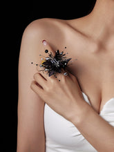 Against a sleek black background, a woman in a white strapless top holds a handcrafted black spiky accessory with colorful pins near her shoulder. She perfectly complements it with the custom symbol CAMELLIA Scorpio Flower Ring.